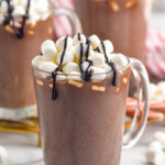 mug of homemade Hot Chocolate topped with marshmallows and chocolate sauce. Two mugs of Hot Chocolate sitting in background, marshmallows sitting beside