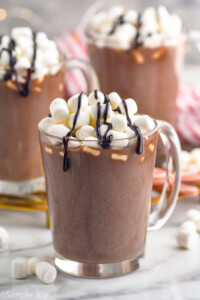 mug of homemade Hot Chocolate topped with marshmallows and chocolate sauce. Two mugs of Hot Chocolate sitting in background, marshmallows sitting beside