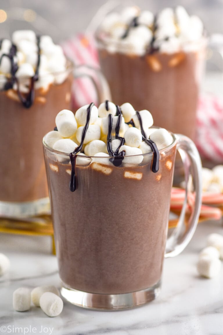 mug of homemade Hot Chocolate topped with marshmallows and chocolate sauce. Two mugs of Hot Chocolate sitting in background, marshmallows sitting beside