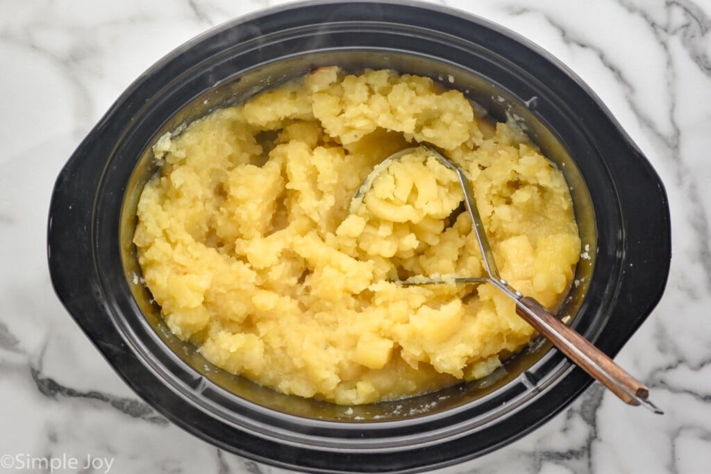 overhead of crockpot of mashed potatoes with potato masher