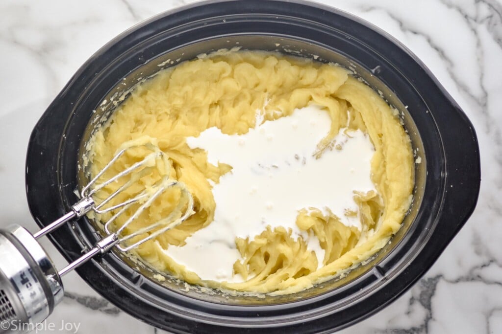 overhead of crockpot of mashed potatoes topped with cream, handheld mixer sitting beside