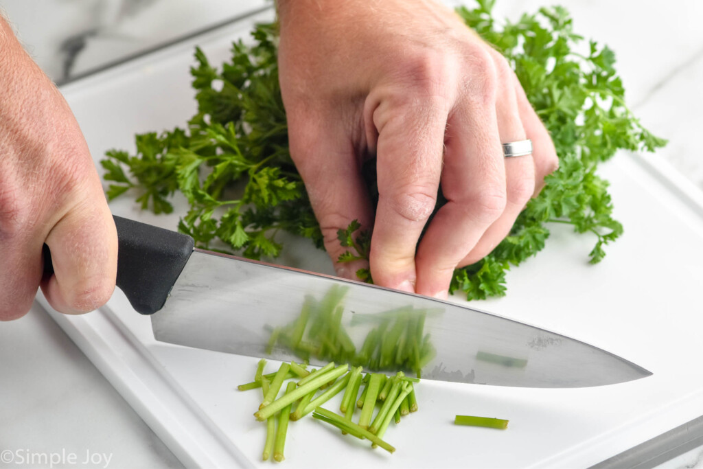 man's hands holding bunch of fresh parsley on a cutting board and sharp knife cutting off stems of parsley to show How to Store Parsley