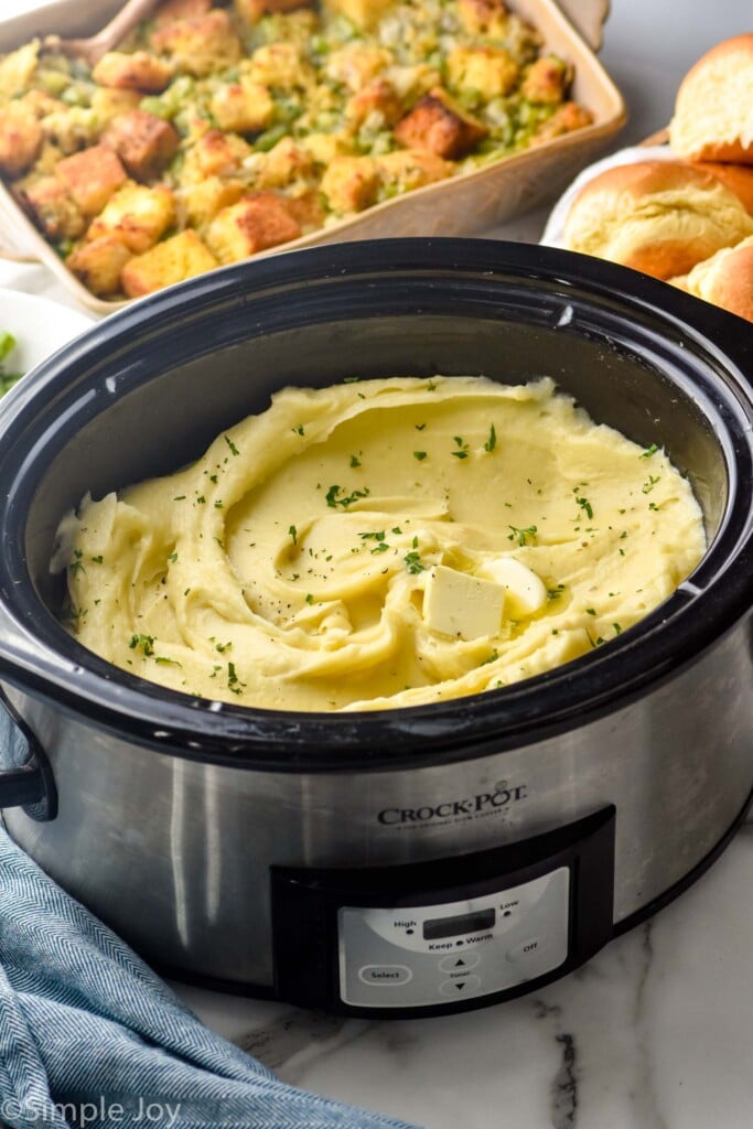 crockpot full of Crockpot Mashed Potatoes topped with butter and fresh parsley. Casserole dish of brussels sprouts casserole sitting in background