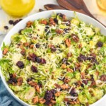 Bowl of Shaved Brussels Sprout Salad topped with shredded parmesan cheese with dressing and spoon for serving sitting in background