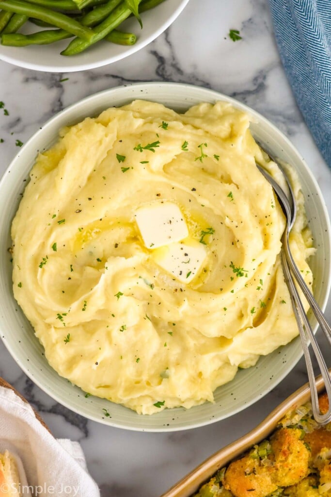 overhead of bowl of Crockpot Mashed Potatoes topped with butter and fresh parsley with two spoons for serving