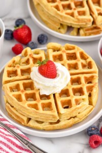 plate of homemade waffles topped with whipped cream and a strawberry. Fresh berries and plate of waffles sitting in background