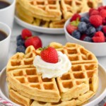 Plate of homemade waffles topped with whipped cream and a strawberry. Bowl of fresh fruit, plate of waffles, and two mugs of coffee sitting in background