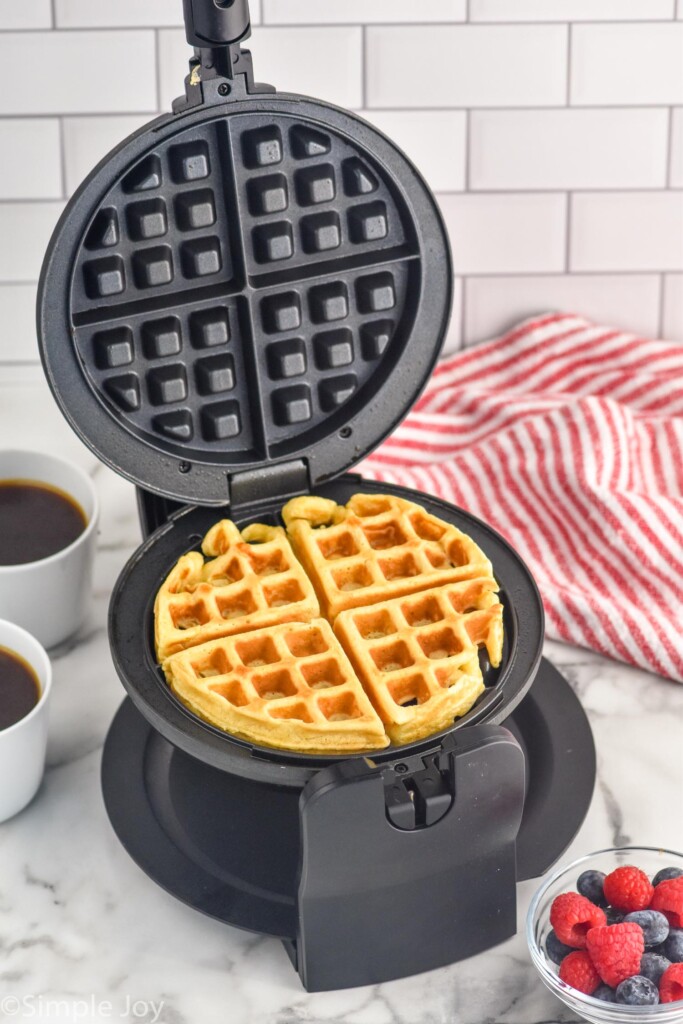 Waffle maker with homemade waffle inside, bowl of fresh berries and two mugs of black coffee sitting beside