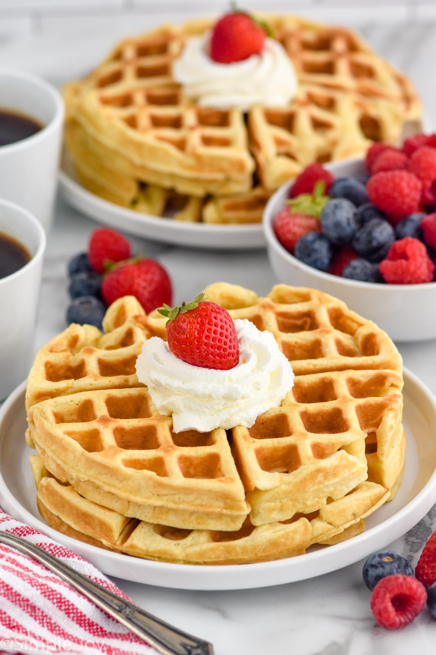 Plate of homemade waffles topped with whipped cream and a strawberry. Bowl of fresh fruit, plate of waffles, and two mugs of coffee sitting in background