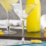 Pinterest graphic for Limoncello Martini. Image shows man's hand pouring cocktail shaker of limoncello martini ingredients into a martini glass. Bottle of limoncello and glass of limoncello martini sitting in background. Says "limoncello martini simplejoy.com"