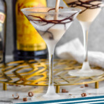 Pinterest graphic for Mudslide Cocktail. Image shows man's hand pouring cocktail shaker of mudslide ingredients into a martini glass with chocolate drizzle. Mudslide cocktail and bottle of Kahlua sitting in background. Text says "mudslide cocktail simplejoy.com"