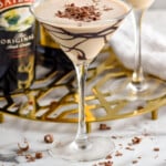 Pinterest graphic for mudslide. Image shows martini glass of Mudslide topped with chocolate shavings. Glass of mudslide, bottle of bailey's irish cream, and bottle of kahlua sitting in background. Says "super easy mudslide cocktail simplejoy.com"