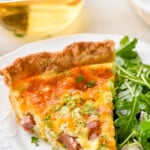 Pinterest graphic for quiche recipe. Text says "The best simple quiche recipe simplejoy.com" Image shows a piece of homemade quiche on a plate with arugula salad sitting beside.