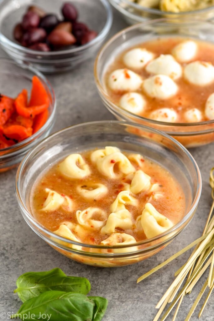 bowl of tortellini in italian dressing with other ingredients sitting behind to make Antipasto Skewers