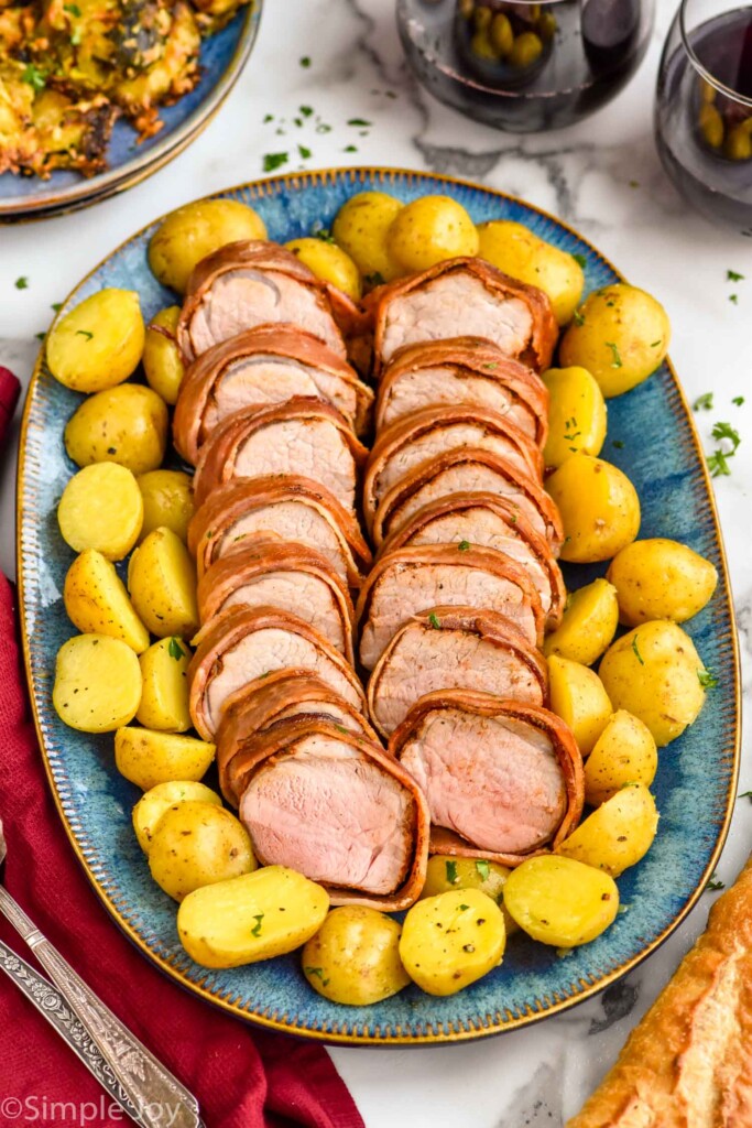 Platter of sliced Bacon Wrapped Pork Tenderloin and potatoes with glasses of wine beside.