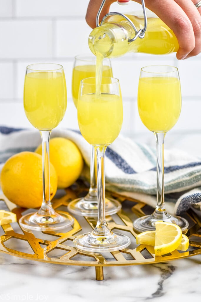 Side view of person's hand pouring Limoncello into wine glasses. Lemons and lemon slices beside.