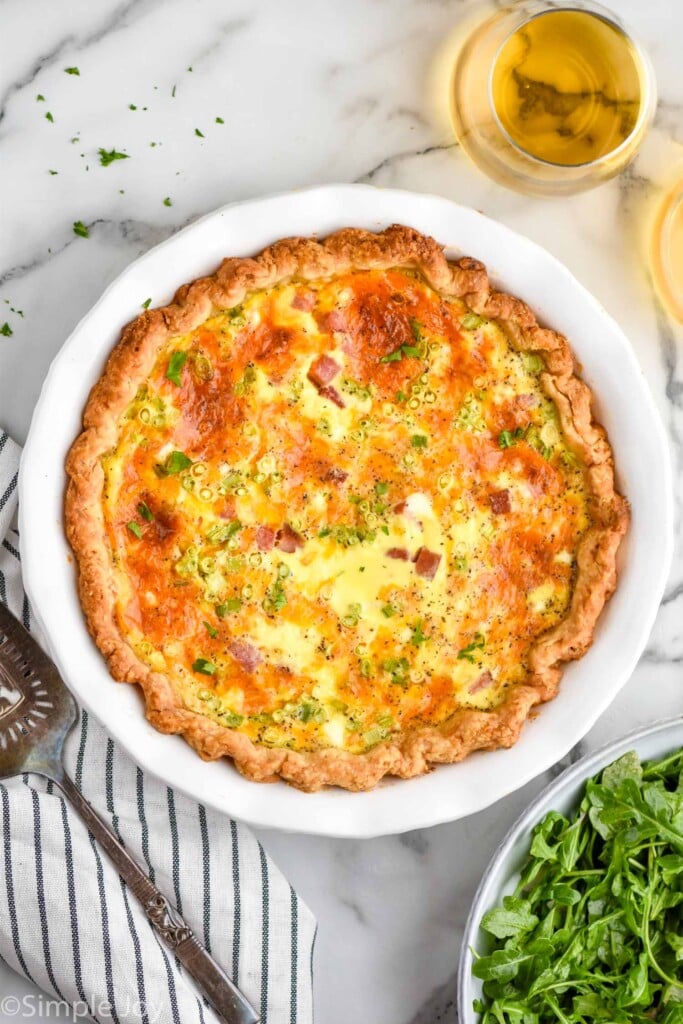 overhead of pie plate of quiche with bowl of arugula salad and two glasses of white wine sitting beside