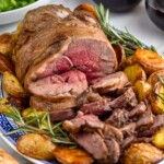 platter of Boneless Leg of Lamb, potatoes, and rosemary sprigs. Two glasses of red wine and bowl of salad sitting in background