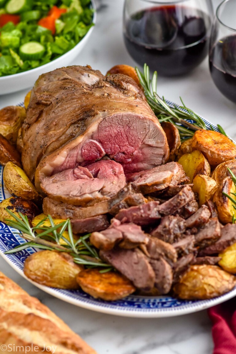 platter of Boneless Leg of Lamb, potatoes, and rosemary sprigs. Two glasses of red wine and bowl of salad sitting in background