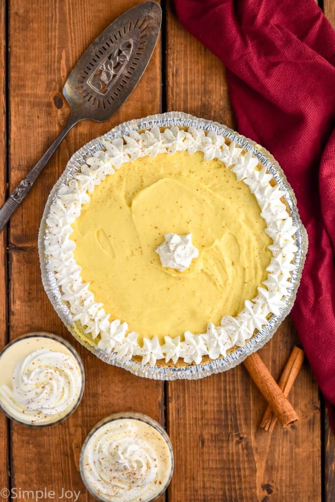 overhead of an eggnog pie topped with whipped cream with two glasses of eggnog, cinnamon sticks, and a spatula for serving sitting beside.