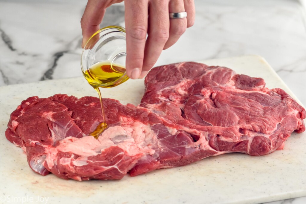 man's hand pouring small cup of olive oil over a Boneless Leg of Lamb sitting on a cutting board