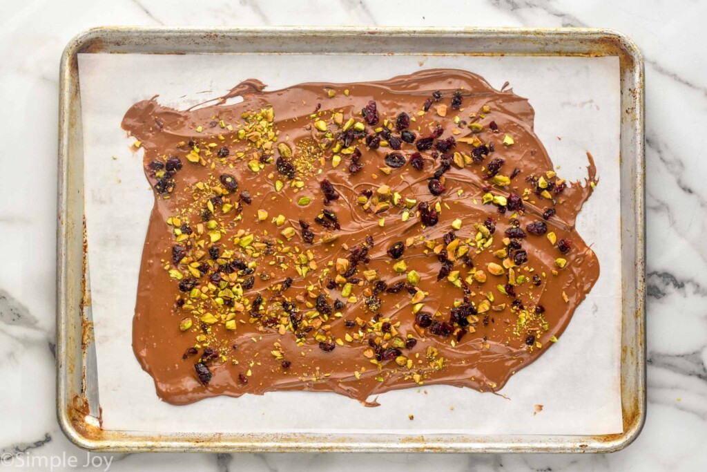 overhead of melted chocolate topped with dried cranberries and nuts on a parchment line rimmed baking sheet to make Chocolate Bark