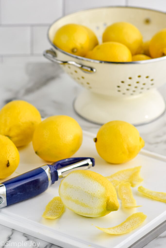 Lemons on cutting board with peeler next to peeled lemon. Colander of more lemons beside for Limoncello recipe.