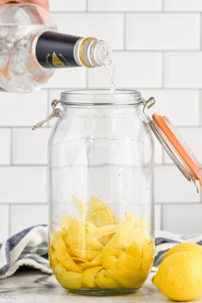 Side view of handle of liqueur being poured into jar of lemon peels for Limoncello recipe. Lemons beside jar.