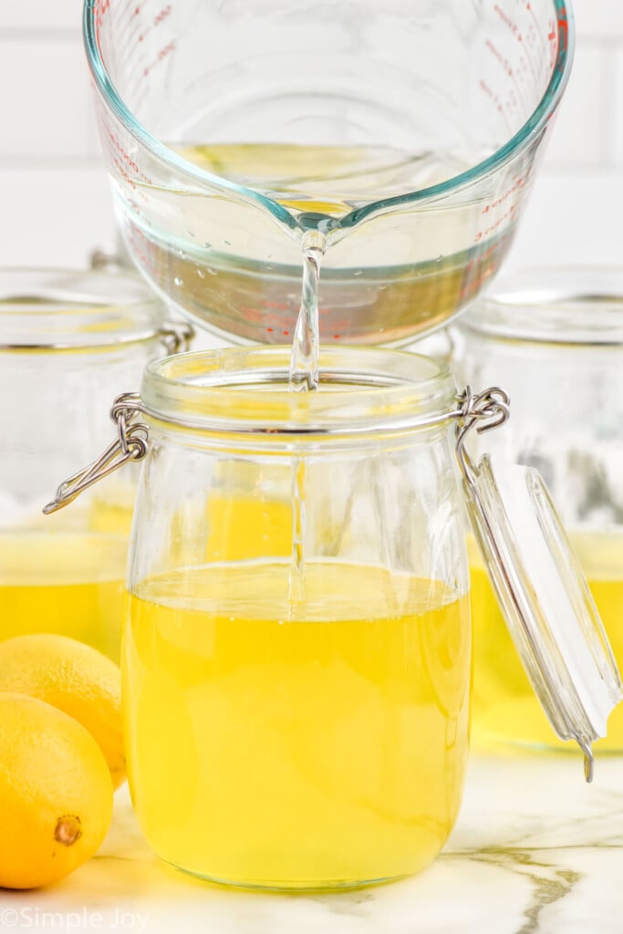 Side view of measuring cup of liquid being poured into a jar of other ingredients for Limoncello recipe. Lemons beside.