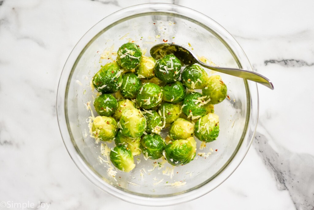 Overhead view of mixing bowl of brussels sprouts with seasoning and shredded cheese with a spoon for mixing for Smashed Brussel Sprouts recipe.