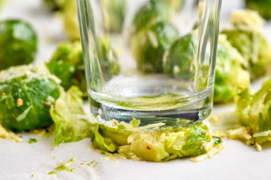 Close up photo of the bottom of a glass smashing a brussels sprout for Smashed Brussel Sprouts recipe. Other brussels sprouts with seasoning and cheese beside.