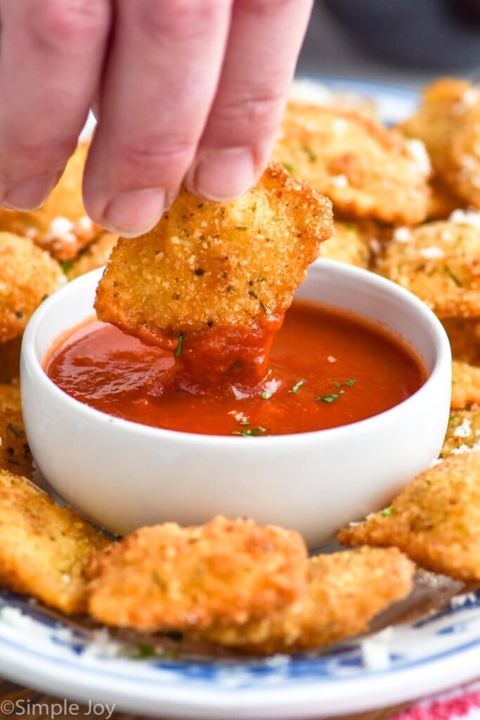 person's hand dipping fried ravioli into small bowl of marinara sauce with fried ravioli sitting on plate.