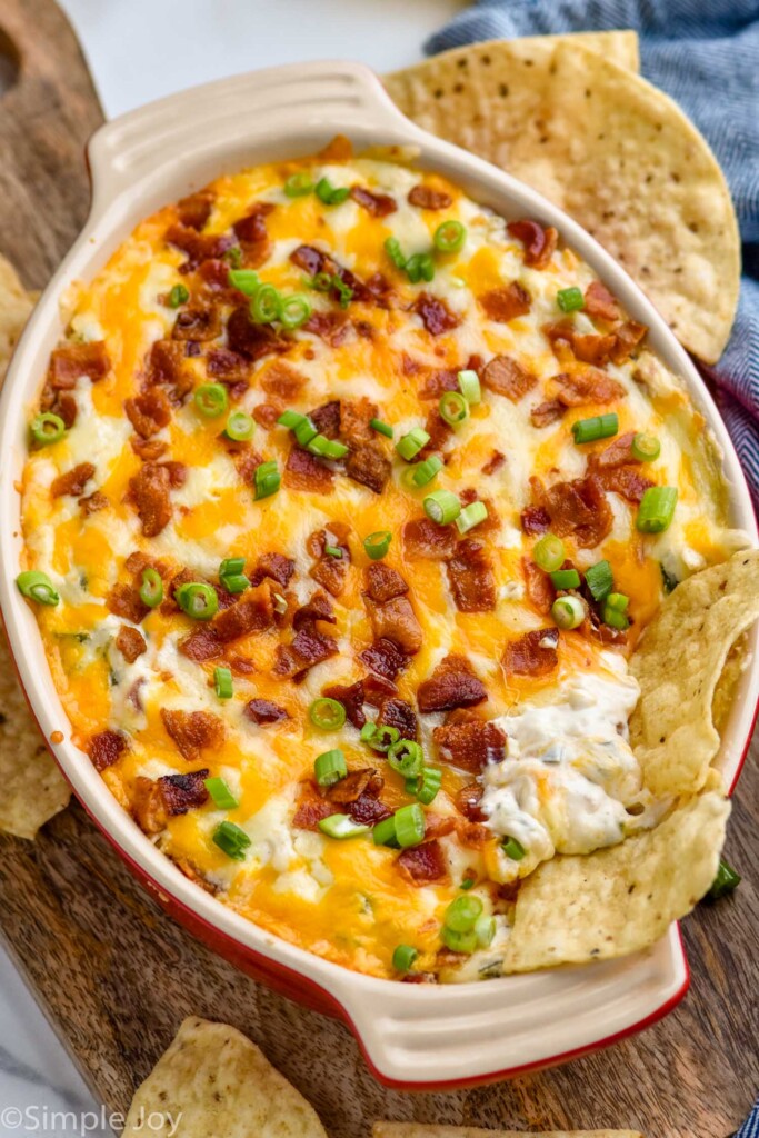 dish of Jalapeño Popper Dip topped with diced bacon and green onion. Tortilla chips sitting in dip and surrounding dish