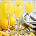 Side view of wine glasses with Limoncello. Jug of Limoncello behind with lemons and lemon slices beside on platter.