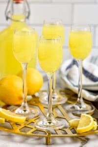 Side view of wine glasses with Limoncello. Jug of Limoncello behind with lemons and lemon slices beside on platter.