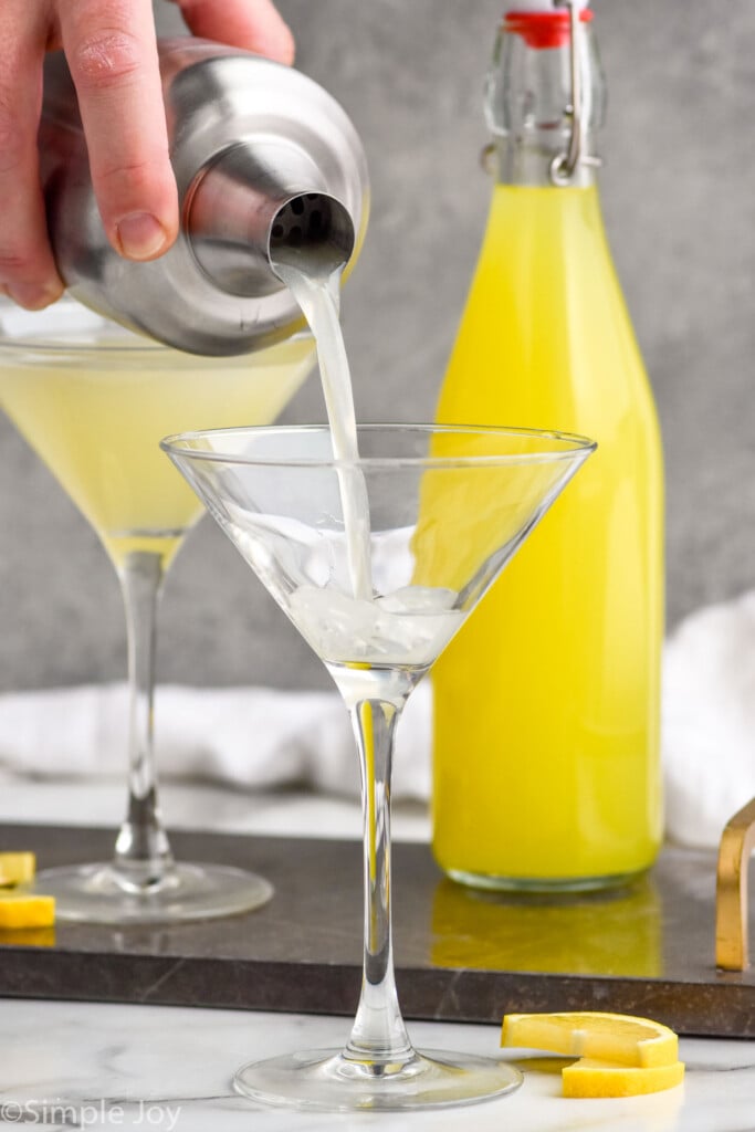 man's hand pouring cocktail shaker of limoncello martini ingredients into a martini glass. Bottle of limoncello and glass of limoncello martini sitting in background