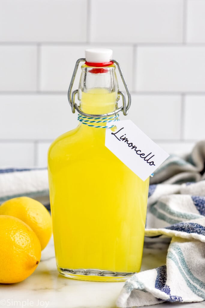 Glass jug of Limoncello with tag that says "Limoncello." Lemons and hand towel beside.