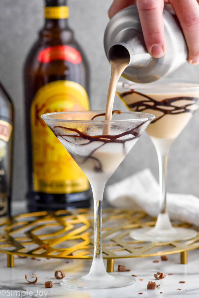 man's hand pouring cocktail shaker of mudslide ingredients into a martini glass with chocolate drizzle. Mudslide cocktail and bottle of Kahlua sitting in background.