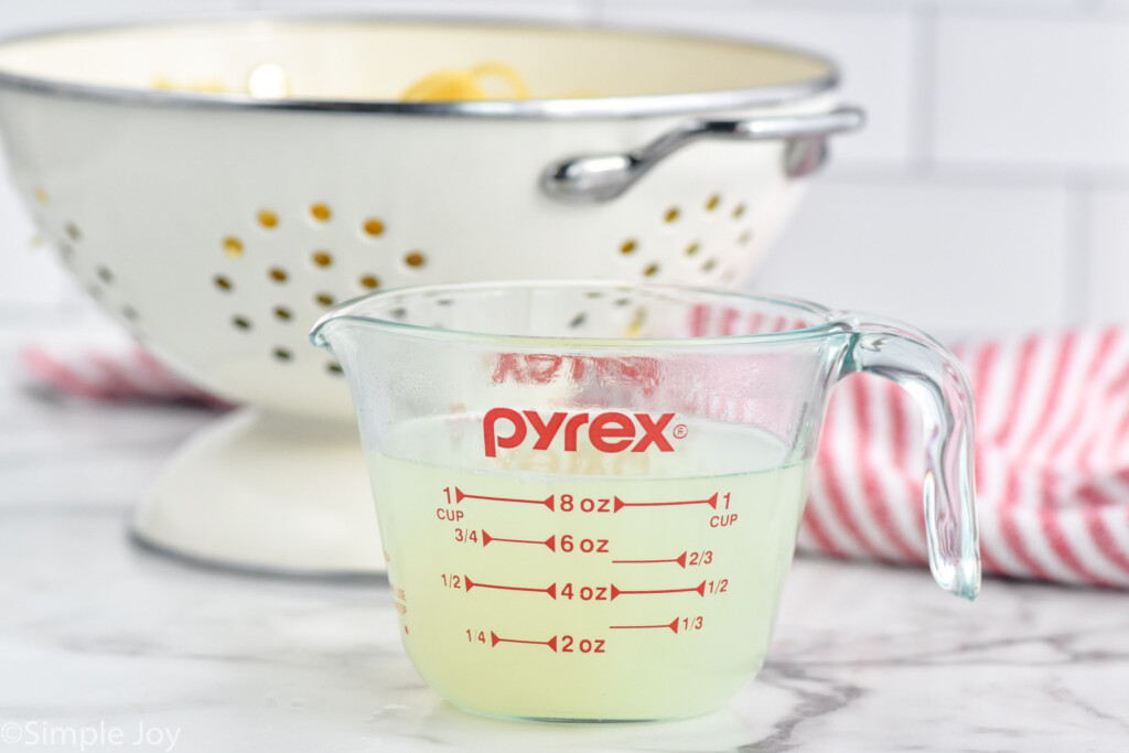 glass measuring cup of reserved pasta water with colander of cooked pasta sitting in background