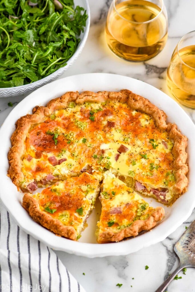 pie plate of homemade quiche with bowl of arugula salad and two glasses of white wine sitting in background