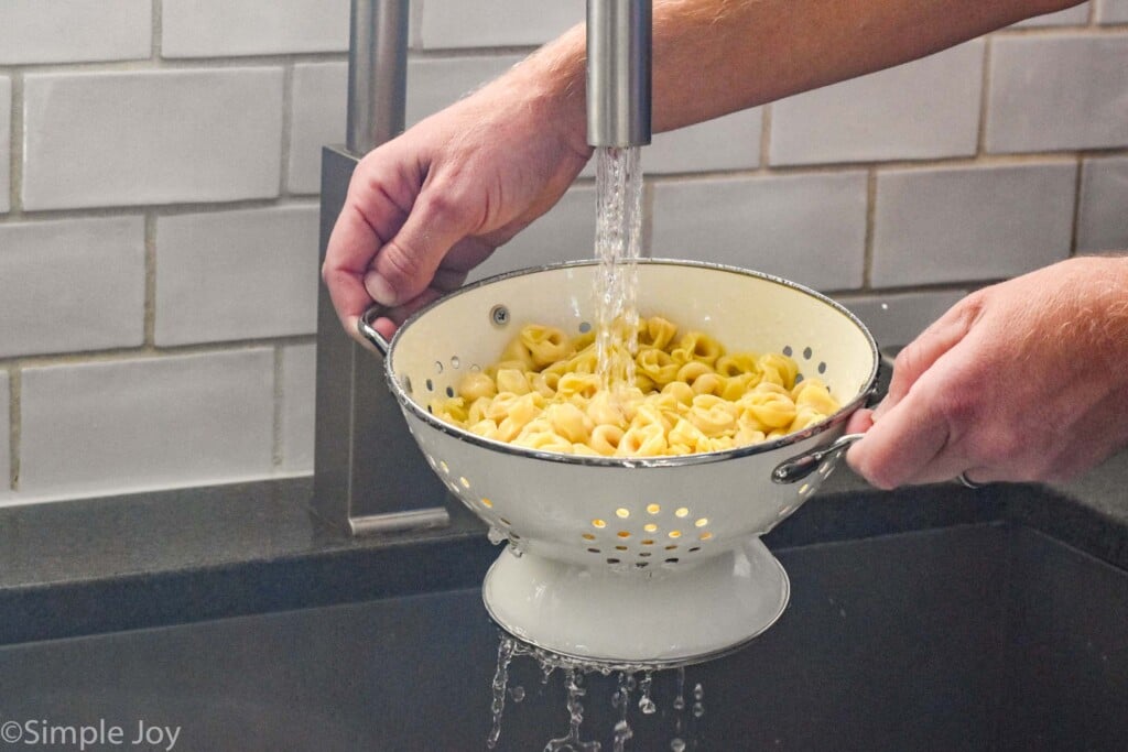 person holding colander of cooked pasta over sink with water running