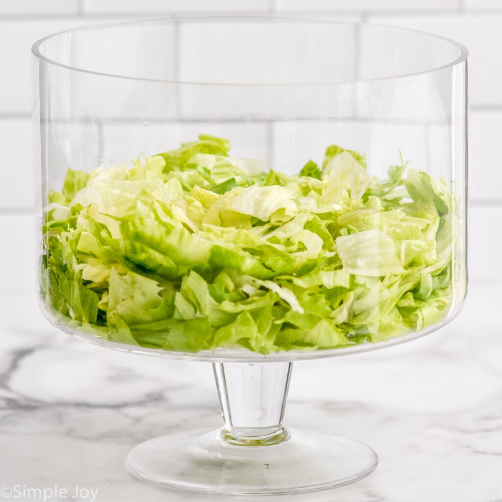 Glass trifle dish of iceberg lettuce to show how to make Seven Layer Salad