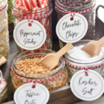 Pinterest graphic for Hot Chocolate Bar. Image shows labeled jars of toppings. Toppings include peppermint sticks, chocolate chips, toffee bits, kosher salt, bottle of caramel sauce, and bottle of chocolate sauce. Says "the best hot chocolate bar set up simplejoy.com"