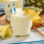 Pinterest graphic for Pineapple Smoothie. Image shows two glasses of Pineapple Smoothie with straws and a pineapple wedge on the rim. Fresh pineapple sitting behind. Says "Pineapple Smoothie simplejoy.com"