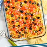 Pinterest graphic for Seven layer dip recipe. Image is overhead view of glass dish of Seven layer dip topped with diced tomatoes, green onions, and black olives. Chips and bottle opener beside. Text says, "Seven layer dip simplejoy.com"