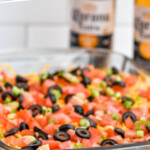 Pinterest graphic for seven layer dip recipe. Text says, "the best seven layer dip simplejoy.com." Image shows side view of a glass dish of seven layer dip topped with cheese, tomatoes, black olives, and green onions. Chips and bottles of beer beside.