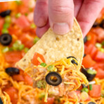 Pinterest graphic for seven layer dip recipe. Text says, "the best seven layer dip simplejoy.com." Close up photo of person's hand holding chip topped with seven layer dip with cheese, tomatoes, green onions, and black olives over glass dish of seven layer dip.