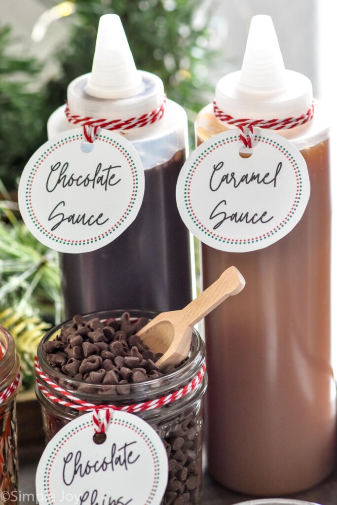 bottles of chocolate sauce and caramel sauce with jar of mini chocolate chips with wooden spoon sitting in front to make a Hot Chocolate Bar