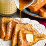 cup of maple syrup pouring onto a plate of pieces Eggnog French Toast topped with powdered sugar and butter. Plate of Eggnog French Toast and glass of eggnog sitting in background.