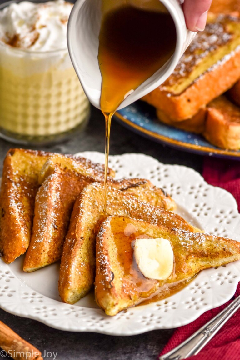 cup of maple syrup pouring onto a plate of pieces Eggnog French Toast topped with powdered sugar and butter. Plate of Eggnog French Toast and glass of eggnog sitting in background.